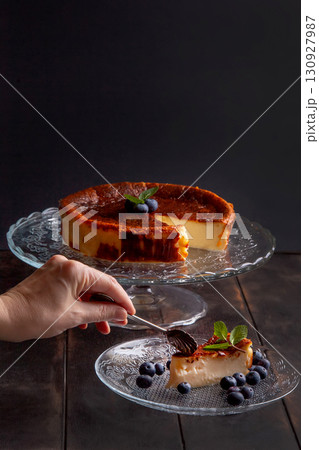 Homemade Basque burnt cheesecake with cream cheese, eggs and cream baked in a crumb crust, with blueberries and mint leaves. Woman breaks off a piece on a plate on dark background Homemade Basque burnt cheesecake with cream cheese, eggs and cream baked in a crumb crust, with blueberries and mint leaves. Woman breaks off a piece on a plate on dark background 130927987