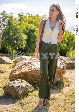 Young woman dressed in white vest and green trousers posing outdoors in Old Money style. Concept of timeless elegance, fashion confidence and classic aesthetic in modern lifestyle. 130928256