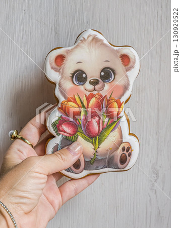 Hand holDecorated gingerbread bear cookie holding flowers in human hand. Cute homemade pastry, sweet festive dessert, creative holiday treat.ding gingerbread cookie shaped like a bear with flowers 130929522