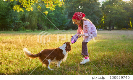 Girl in bandana playing frisbee with dog on meadow in golden light Girl in bandana playing frisbee with dog on meadow in golden light 130929740