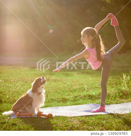 Girl balancing in yoga pose on mat while dog watches under sunlight outdoors 130929765