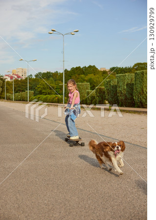 Girl riding skateboard with dog running beside in city park Girl riding skateboard with dog running beside in city park 130929799