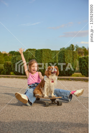 Girl sitting with dog on skateboard in park with joyful expression 130929800