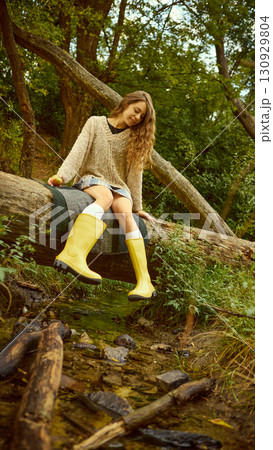 Woman sitting on log above stream in yellow boots with serene smile 130929804