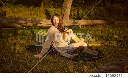 Young woman sitting on blanket in forest with cup smiling warmly in golden light 130929824
