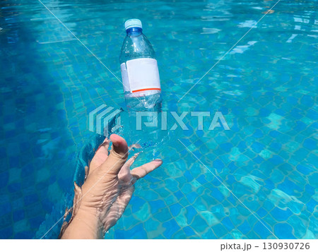 A hand lifts a plastic bottle from clear blue pool water, symbolizing pollution cleanup and environmental awareness. A hand lifts a plastic bottle from clear blue pool water, symbolizing pollution cleanup and environmental awareness. 130930726