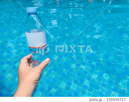 A hand lifts a plastic bottle from clear blue pool water, symbolizing pollution cleanup and environmental awareness. 130930727
