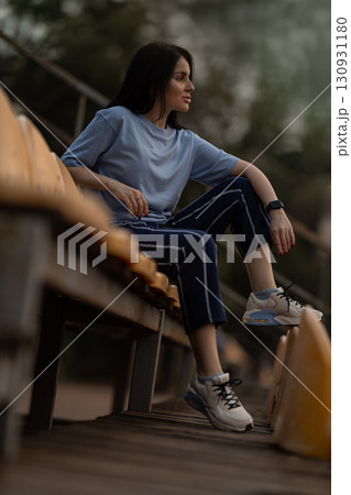 brunette girl sits comfortably on the empty stadium stands wearing a blue t shirt and casual pants. She looks relaxed enjoying the peaceful atmosphere in the venue. brunette girl sits comfortably on the empty stadium stands wearing a blue t shirt and casual pants. She looks relaxed enjoying the peaceful atmosphere in the venue. 130931180