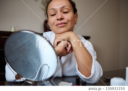 Woman Reflecting in Mirror Wearing White Robe for Skincare Routine Woman Reflecting in Mirror Wearing White Robe for Skincare Routine 130931350