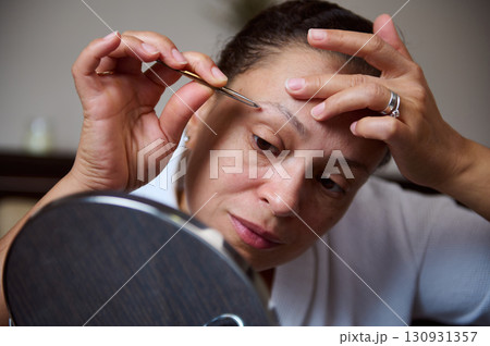 Woman Tweezing Eyebrows in Front of a Mirror at Home Woman Tweezing Eyebrows in Front of a Mirror at Home 130931357