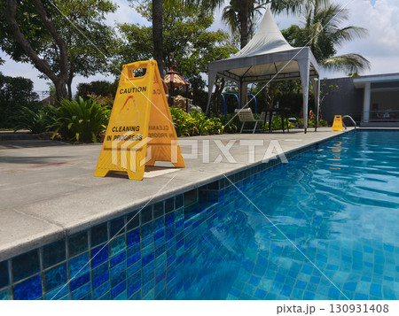 Yellow caution sign reading "Cleaning in Progress" placed near the edge of a swimming pool, emphasizing safety and maintenance in an outdoor area. 130931408