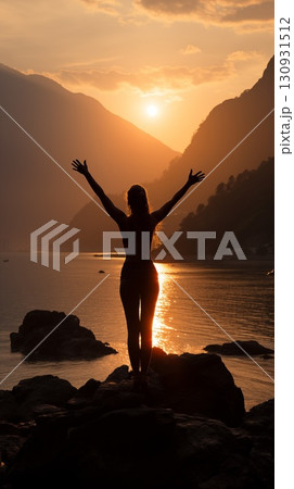 Silhouette of a woman in the sunset, standing on rocks with arms raised, facing a sunset over a calm sea with mountains in the background 130931512