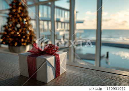 A gift box with a red ribbon stands in a room against the backdrop of a Christmas tree and a panorama of the sea outside the window. Christmas on the coast. Happy New Year. 130931962