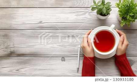 Two hands gently cradle a steaming cup of tea above a wooden table, accompanied by a pen and green plants in the background 130933180
