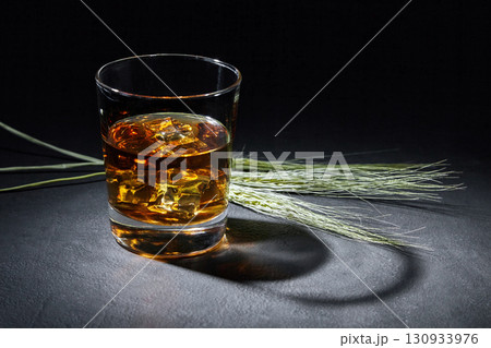 glass of amber whiskey and ears of rye on a black stone table. glass of amber whiskey and ears of rye on a black stone table. 130933976