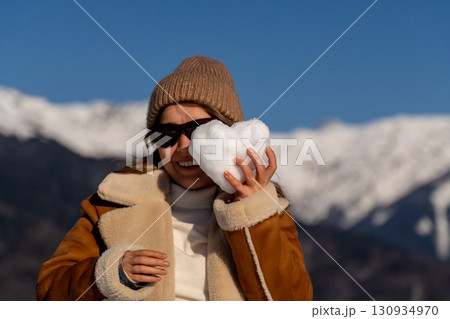 Snow Heart Mountains Winter: Woman playfully holds heart-shaped snowball, sunny day, joyful winter activity. 130934970