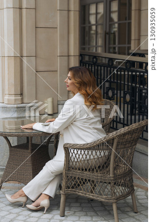 Woman White Suit Outdoor Cafe - Stylish woman in a white suit sitting on a patio chair at an outdoor cafe. 130935059