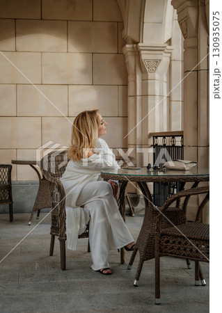Woman White Outfit Patio Cafe - Sitting alone at a table on a patio outside a cafe, a woman in a white outfit enjoys a relaxing moment. Woman White Outfit Patio Cafe - Sitting alone at a table on a patio outside a cafe, a woman in a white outfit enjoys a relaxing moment. 130935075