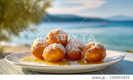 A plate of delicious Greek loukoumades, or honey doughnuts, is presented outdoors with sea view in the background. The warm, fried dough balls are drenched in honey and sprinkled with powdered sugar 130935435