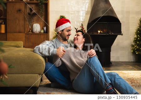 Merry Christmas. Happy young adult couple hugging bonding near Christmas tree and fireplace. Happy family man woman at home. People together celebrating Christmas enjoying winter holidays at home 130935445