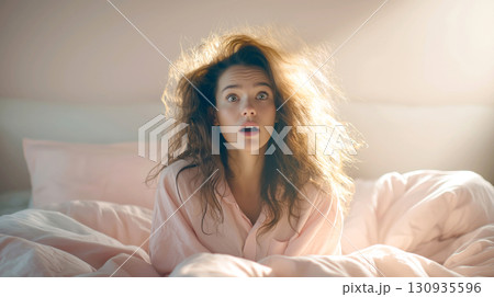 A young woman with wild, messy bedhead hair wakes up with shocked, surprised expression. The image captures humorous and relatable moment of morning confusion, with warm, soft light flooding bedroom 130935596
