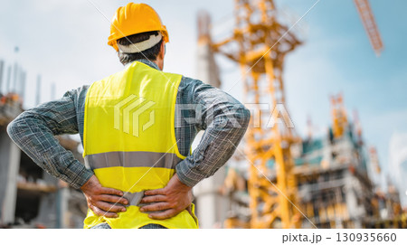 A construction worker, seen from behind, wearing a hard hat and high-visibility vest, holding his lower back in pain while observing a bustling construction site with cranes and buildings A construction worker, seen from behind, wearing a hard hat and high-visibility vest, holding his lower back in pain while observing a bustling construction site with cranes and buildings 130935660