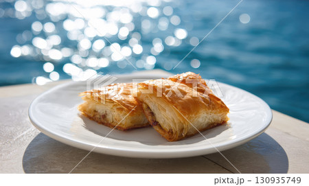 A close-up of two pieces of golden-brown baklava, a traditional Greek pastry, served on a clean white plate. The background features blue ocean with sun glare, creating vacation, summer atmosphere A close-up of two pieces of golden-brown baklava, a traditional Greek pastry, served on a clean white plate. The background features blue ocean with sun glare, creating vacation, summer atmosphere 130935749