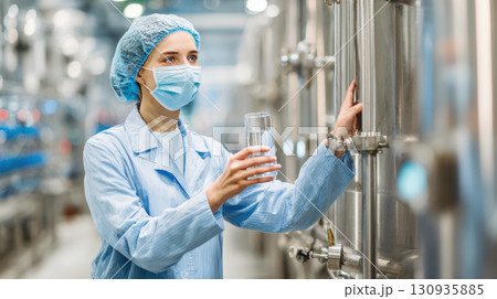 quality control technician in sterile uniform, holding glass of water for inspection in beverage production facility. Image represents hygiene, safety, quality assurance, drink industry 130935885