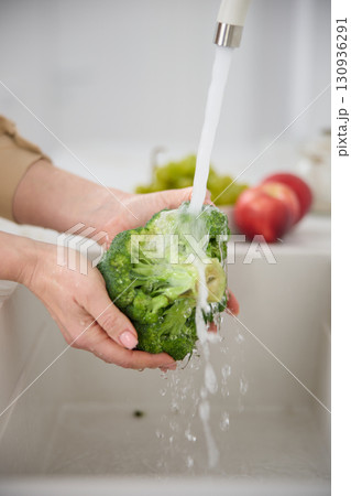 Close up view of a woman washes broccoli under the pressure of flowing water in the white kitchen, tomatoes and green grapes in the background Close up view of a woman washes broccoli under the pressure of flowing water in the white kitchen, tomatoes and green grapes in the background 130936291