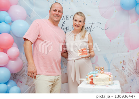 Smiling couple standing with cake at gender reveal party. Festive balloons in pink and blue colors, lifestyle editorial family event concept 130936447