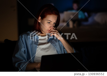 Serious focused woman freelancer working on laptop in dark at home looking at screen.  130936552