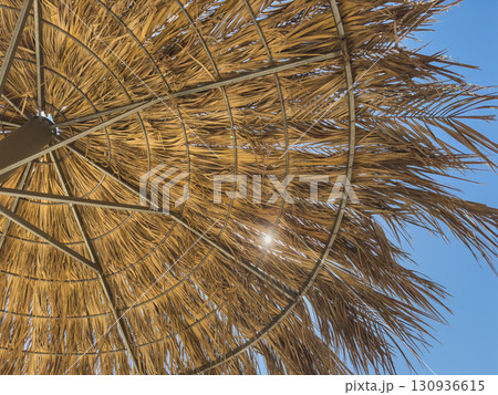 Parasol straw umbrella with sunlight rays. Straw umbrella on hot summer day by beach. Sunny blue skies warm weather enjoyed on beach under parasol made of straw. Vacation in tropical countries 130936615