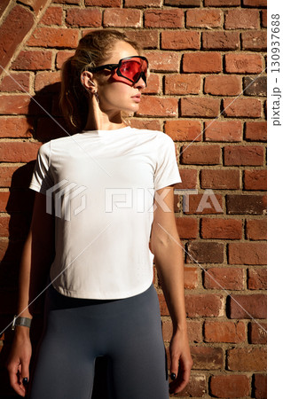 Young woman in sporty attire posing against a brick wall in sunlight 130937688