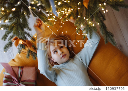 Smiling child top view under Christmas tree with gift box on orange cushion on wooden floor Smiling child top view under Christmas tree with gift box on orange cushion on wooden floor 130937793