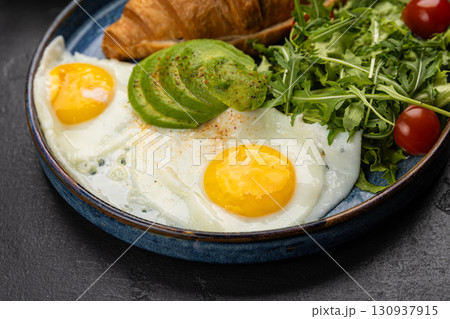 Healthy breakfast with croissant, avocado, sunny side eggs and salad on dark background 130937915