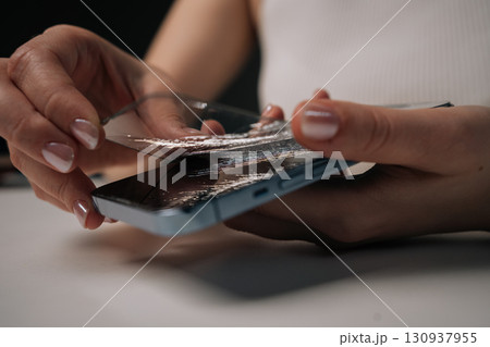 Hands of young unrecognizable woman removing broken tempered glass screen protector from mobile phone, showing importance of screen protection and potential damage it prevents. 130937955