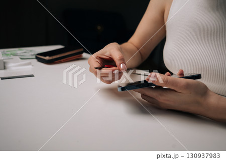 Young woman removing broken tempered glass screen protector from mobile phone with plastic opening tool, sitting at white table, focusing on repair process. Concept of mobile phone service. 130937983