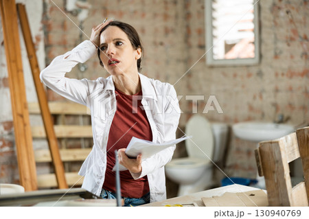 Confused female designer standing with papers in apartment during renovation 130940769