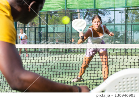 Female player playing padel in a padel court outdoor behind net 130940903