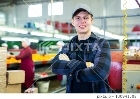 Positive male employee in a warehouse next to industrial modern tech production sorting line of citrus fruits in packing plant. Positive male employee in a warehouse next to industrial modern tech production sorting line of citrus fruits in packing plant. 130941308