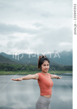 Woman in activewear doing yoga by a lake with mountains in the background, under a cloudy sky. 130945932