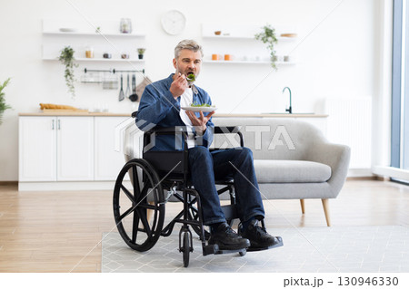 Middle-aged man sitting in wheelchair enjoying a salad in a bright living room. Scene emphasizes inclusion, accessibility, and healthy eating lifestyle. White kitchen with modern furniture Middle-aged man sitting in wheelchair enjoying a salad in a bright living room. Scene emphasizes inclusion, accessibility, and healthy eating lifestyle. White kitchen with modern furniture 130946330