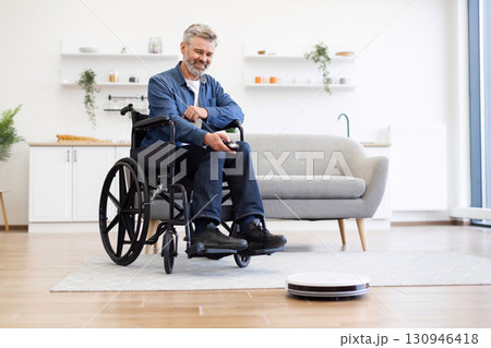 Senior man seated in wheelchair operating robot vacuum cleaner in bright tidy living room. Demonstrates accessibility, technology use, and independence in daily tasks. Senior man seated in wheelchair operating robot vacuum cleaner in bright tidy living room. Demonstrates accessibility, technology use, and independence in daily tasks. 130946418