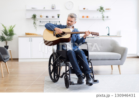 Mature white man playing acoustic guitar in wheelchair in modern living space, expressing music Mature white man playing acoustic guitar in wheelchair in modern living space, expressing music 130946431