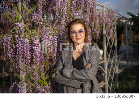 Woman Wisteria Garden Portrait - A woman in a grey jacket stands in front of a trellis of wisteria flowers in a garden. Woman Wisteria Garden Portrait - A woman in a grey jacket stands in front of a trellis of wisteria flowers in a garden. 130950007