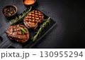 Grilled beef steaks with rosemary, salt, and pepper on wooden board. Juicy barbecue meat with spices. Delicious cooked beef fillet on dark rustic background. 130955294