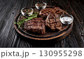 Grilled beef steaks with rosemary, salt, and pepper on wooden board. Juicy barbecue meat with spices. Delicious cooked beef fillet on dark rustic background. 130955298