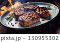 Grilled beef steaks with rosemary, salt, and pepper on wooden board. Juicy barbecue meat with spices. Delicious cooked beef fillet on dark rustic background. 130955302