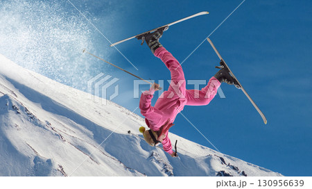 Female skier in pink suit flipping upside down mid-air over snowy slope with dramatic energy. Hand made collage 130956639