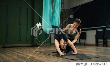 Female trainer assisting young woman in aerial yoga inversion using hammock fabric 130957817